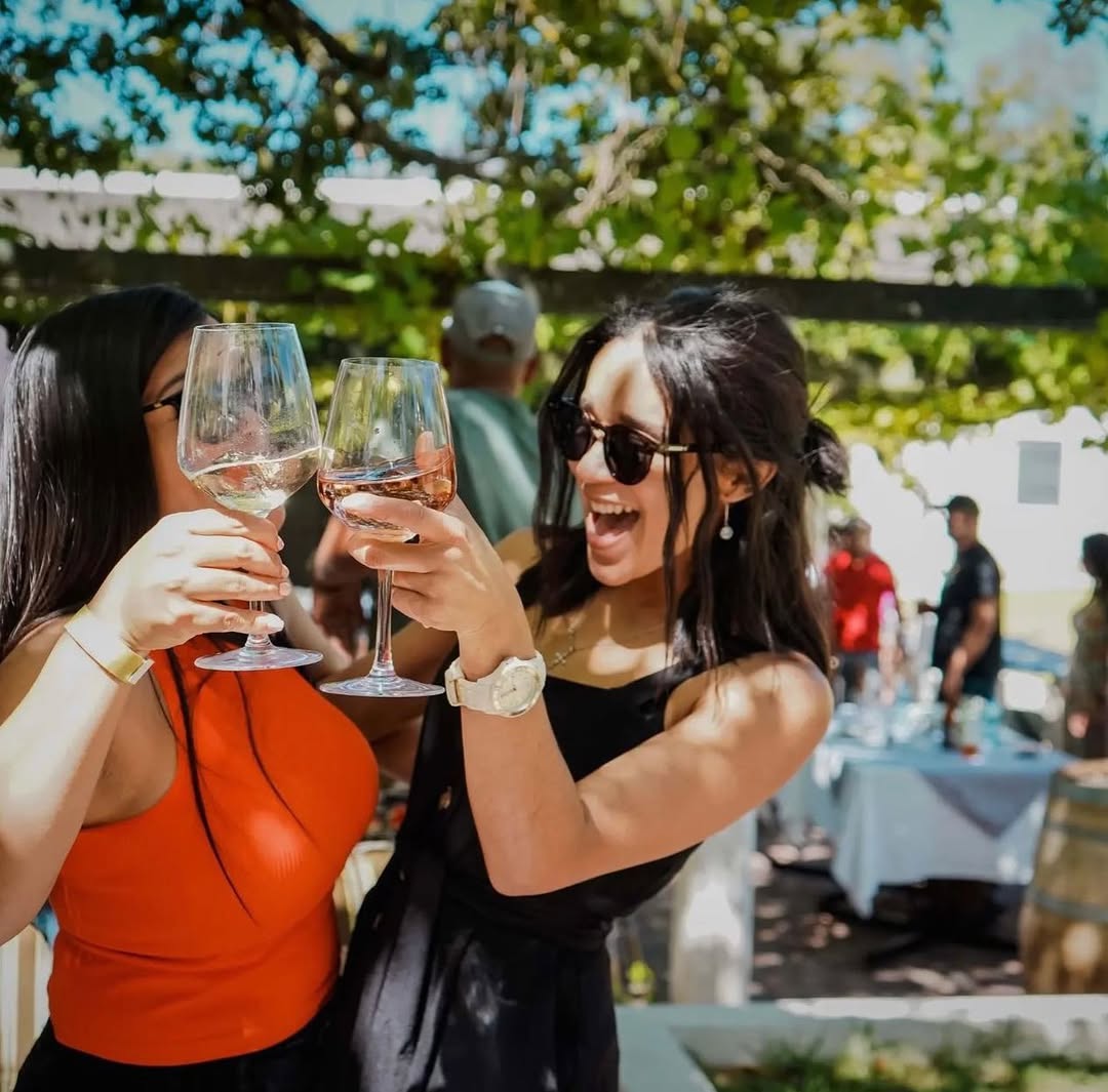 Two women laughing and toasting glasses of wine at an outdoor event under a vine pergola